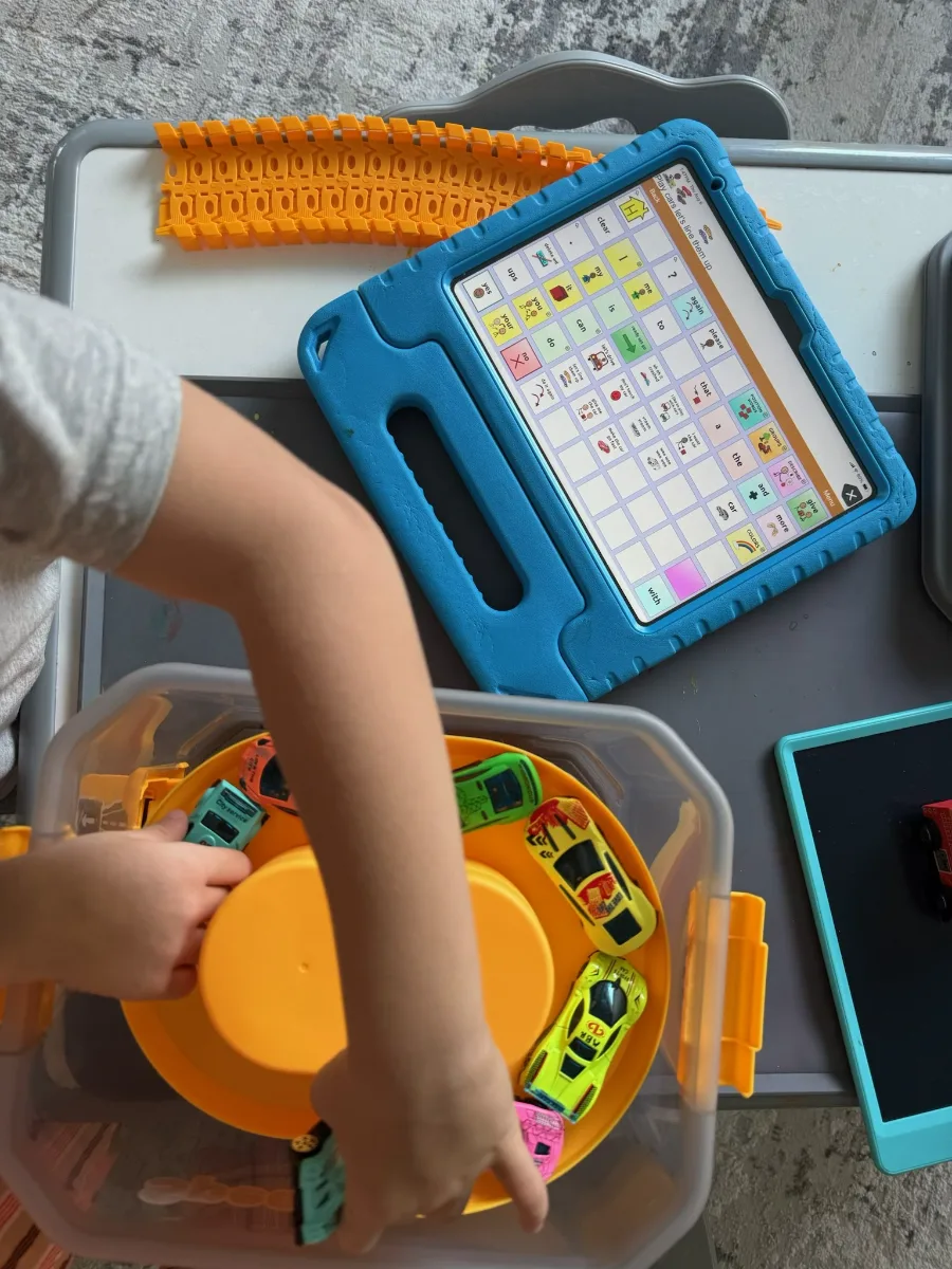 A child playing with toy cars while using an AAC communication device on a tablet