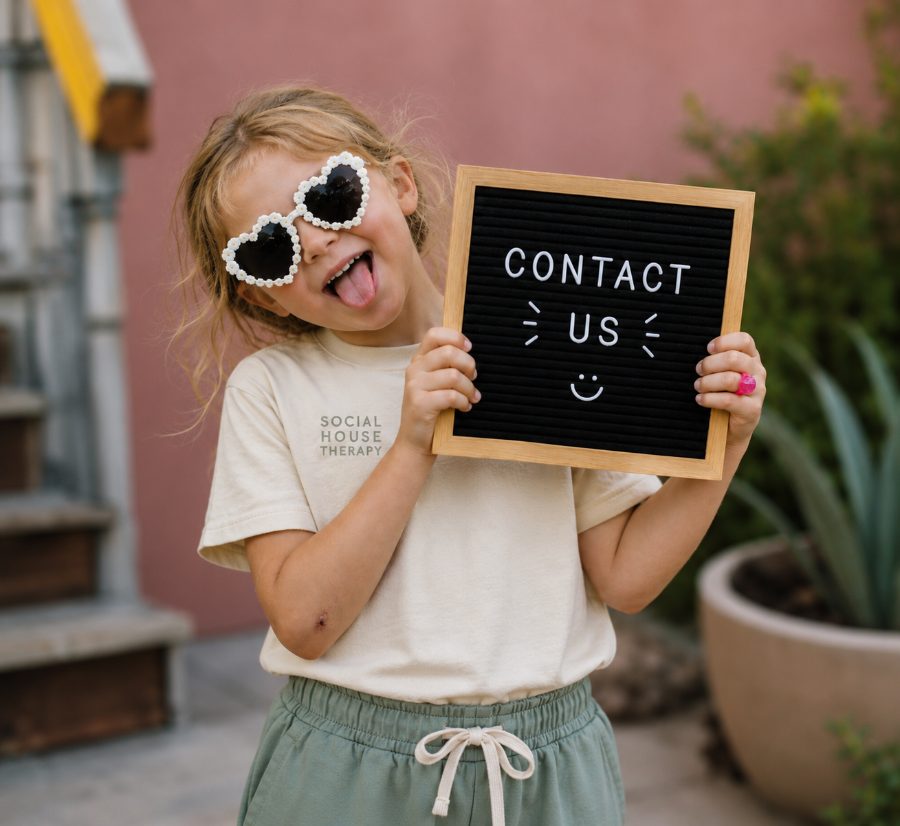 A smiling child in heart-shaped sunglasses and a Social House Therapy shirt holding a letter board that says Contact Us