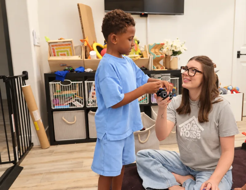 Madison Jeffery, speech-language pathologist, sitting on a playroom floor and smiling up at a young boy as he hands her a toy monster truck