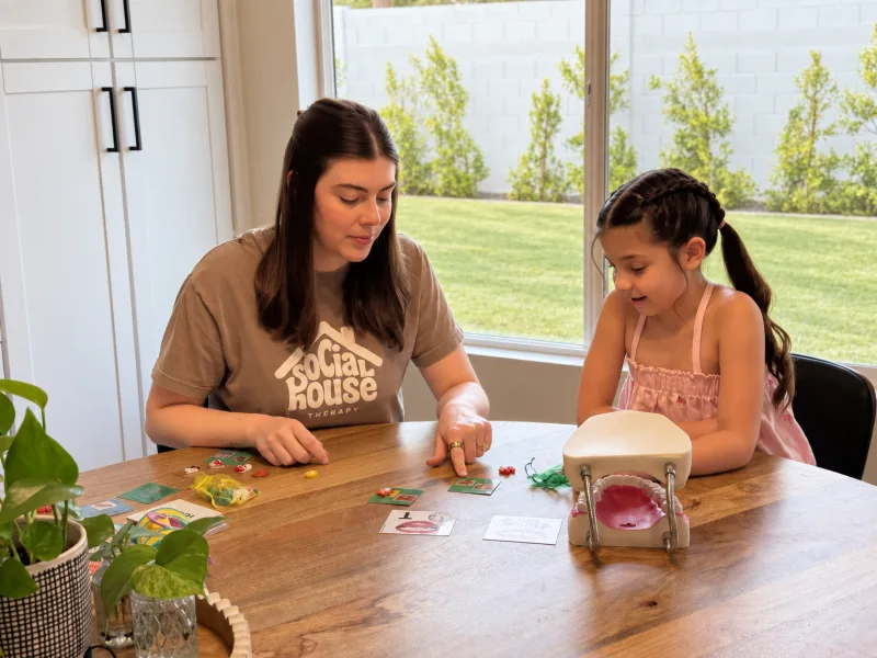 Madison Jeffery and a young girl sitting at a kitchen table working through speech sound cards, with a model of the mouth and teeth between them