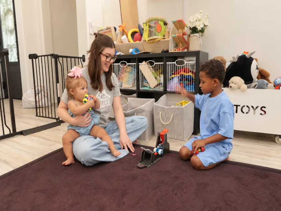 Madison Jeffery sitting on a playroom rug with a baby on her lap while a young boy kneels across from her playing with a toy car ramp