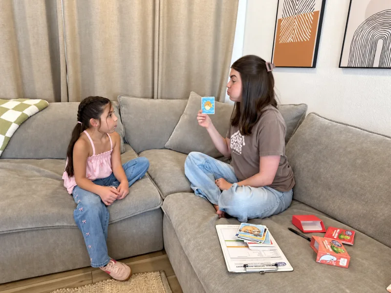 Madison Jeffery sitting cross-legged on a living room couch holding up a picture card for a young girl during a speech therapy session